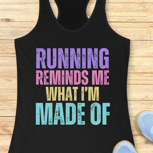 A black racerback tank top laid flat on a light wood background, featuring the text 'RUNNING REMINDS ME WHAT I'M MADE OF' in distressed purple, pink, yellow, and light blue lettering. Partial blue sneakers are visible at the bottom right as styling props.