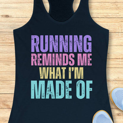 A navy racerback tank top laid flat on a light wood floor, featuring the text 'RUNNING REMINDS ME WHAT I'M MADE OF' in distressed purple, pink, yellow, and light blue lettering. Part of a white sneaker is visible in the bottom right corner.