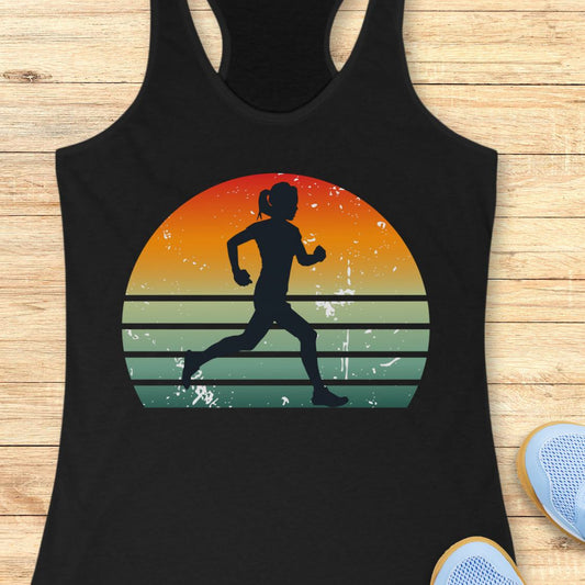 A black racerback tank top laid flat on a light wood surface, featuring a large retro sunset graphic with a black silhouette of a running woman. Part of a blue shoe is visible in the bottom right corner.