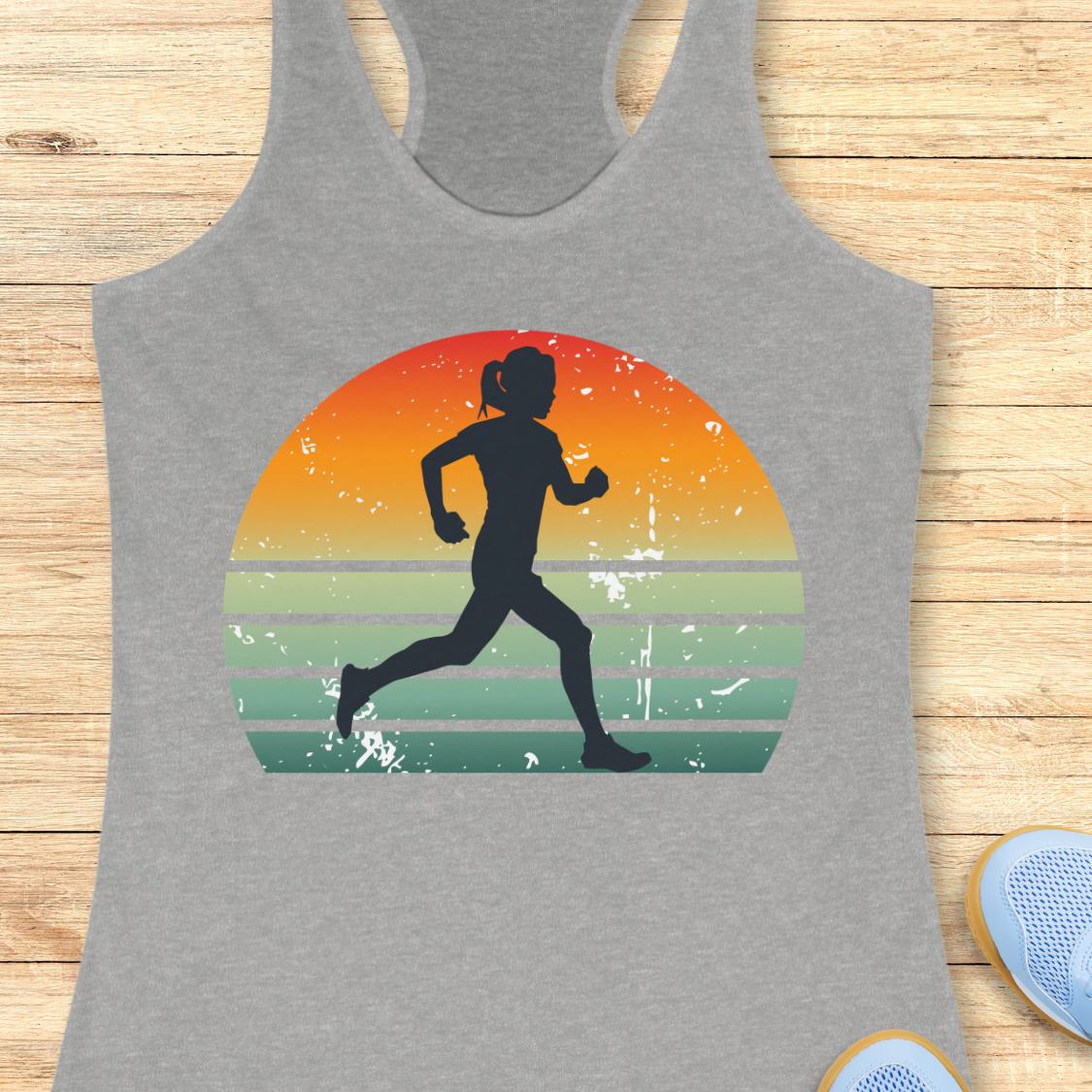 A heather gray racerback tank top laid flat on a light wood surface, featuring a large front graphic of a black running silhouette inside a retro sunset circle with orange, yellow, and green stripes. Partial blue shoes are visible at the bottom right edge.