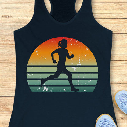 A navy racerback tank top laid flat on a light wood background, featuring a large retro sunset graphic with a black silhouette of a running woman. Part of a white sneaker is visible at the bottom right.