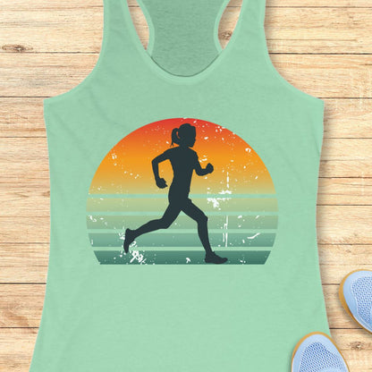 A mint green racerback tank top laid flat on a light wood background, featuring a large graphic of a black running silhouette inside a retro sunset circle with orange, yellow, and teal stripes.
