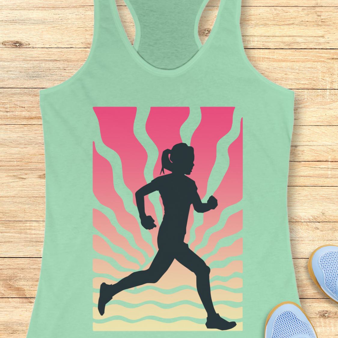 A mint green racerback tank top laid flat on a light wood background, featuring a large graphic print of a black running silhouette against a pink, orange, and yellow wavy sunburst design. Partial blue sneakers are visible at the bottom right edge.
