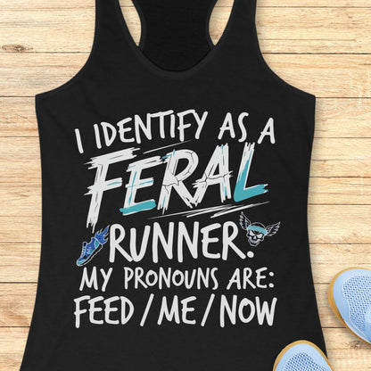 A black racerback tank top laid flat on a light wood surface, printed with the phrase 'I IDENTIFY AS A FERAL RUNNER. MY PRONOUNS ARE: FEED / ME / NOW' in white and light blue text, with a small blue running shoe and skull graphic.