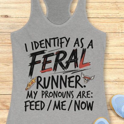 A heather gray racerback tank top laid flat on a light wood surface, printed with the phrase “I IDENTIFY AS A FERAL RUNNER. MY PRONOUNS ARE: FEED / ME / NOW” in black and red lettering, with small graphic accents including a running shoe and a skull wearing a red bandana.