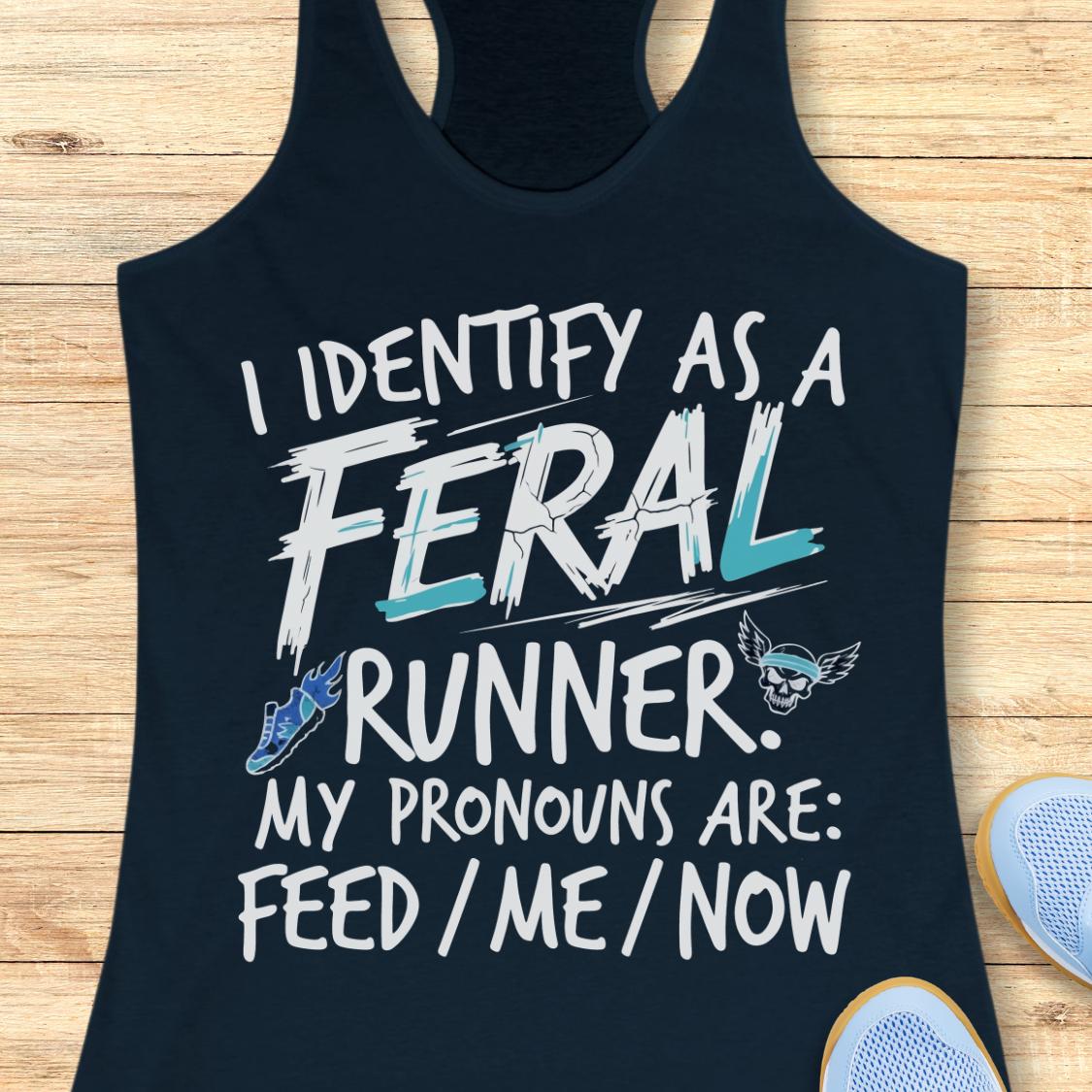 A navy racerback tank top laid flat on a light wood surface, featuring a humorous graphic that reads 'I IDENTIFY AS A FERAL RUNNER. MY PRONOUNS ARE: FEED / ME / NOW' in white and light blue text, with a small blue sneaker and skull-and-wings logo accents.