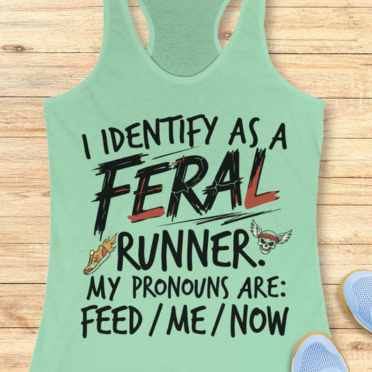 A mint green racerback tank top laid flat on a light wood plank background. The tank top features a graphic print that reads 'I IDENTIFY AS A FERAL RUNNER.