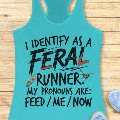 A turquoise racerback tank top laid flat on a light wood surface, printed with the phrase 'I IDENTIFY AS A FERAL RUNNER. MY PRONOUNS ARE: FEED / ME / NOW' in black lettering with red and orange accents, plus small graphic illustrations.