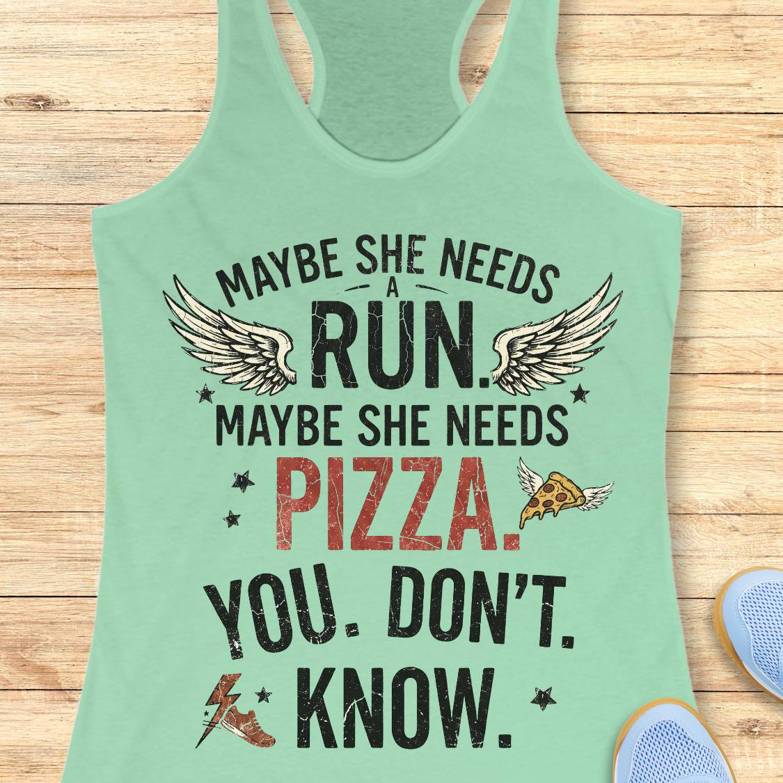 A mint green racerback tank top laid flat on a light wood background, featuring a humorous graphic print that reads 'MAYBE SHE NEEDS A RUN, MAYBE SHE NEEDS PIZZA. YOU DON'T KNOW.