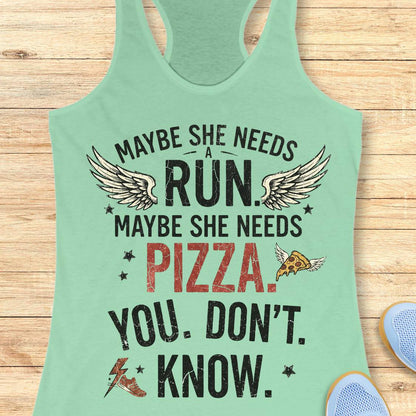 A mint green racerback tank top laid flat on a light wood background, featuring a humorous graphic print that reads 'MAYBE SHE NEEDS A RUN, MAYBE SHE NEEDS PIZZA. YOU DON'T KNOW.