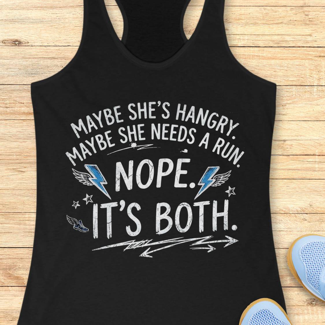 A black racerback tank top laid flat on a light wood surface, featuring a humorous white graphic text that reads 'MAYBE SHE'S HANGRY. MAYBE SHE NEEDS A RUN.