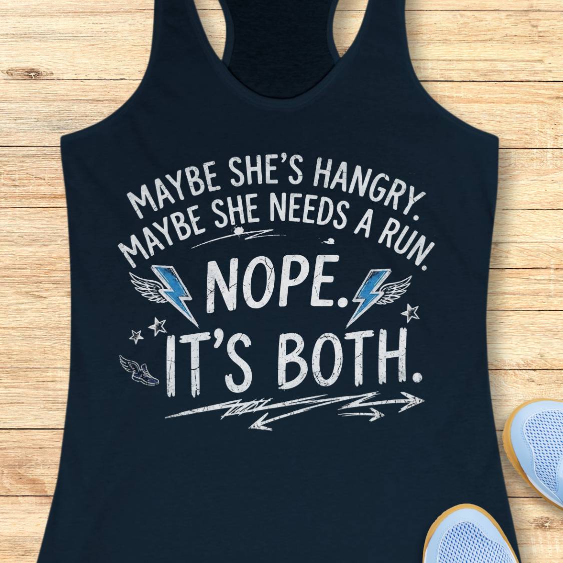 A navy racerback tank top laid flat on a light wood surface, featuring a humorous white graphic text that reads 'MAYBE SHE'S HANGRY. MAYBE SHE NEEDS A RUN.
