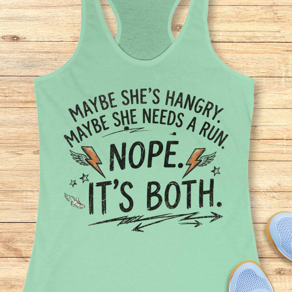 A mint racerback tank top laid flat on a light wood surface, featuring a humorous white graphic text that reads 'MAYBE SHE'S HANGRY. MAYBE SHE NEEDS A RUN.