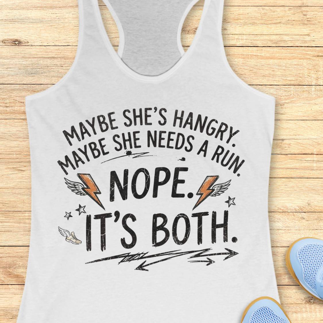 A white racerback tank top with a humorous graphic printed on the front, laid flat on a light wood background. The graphic reads 'MAYBE SHE'S HANGRY.