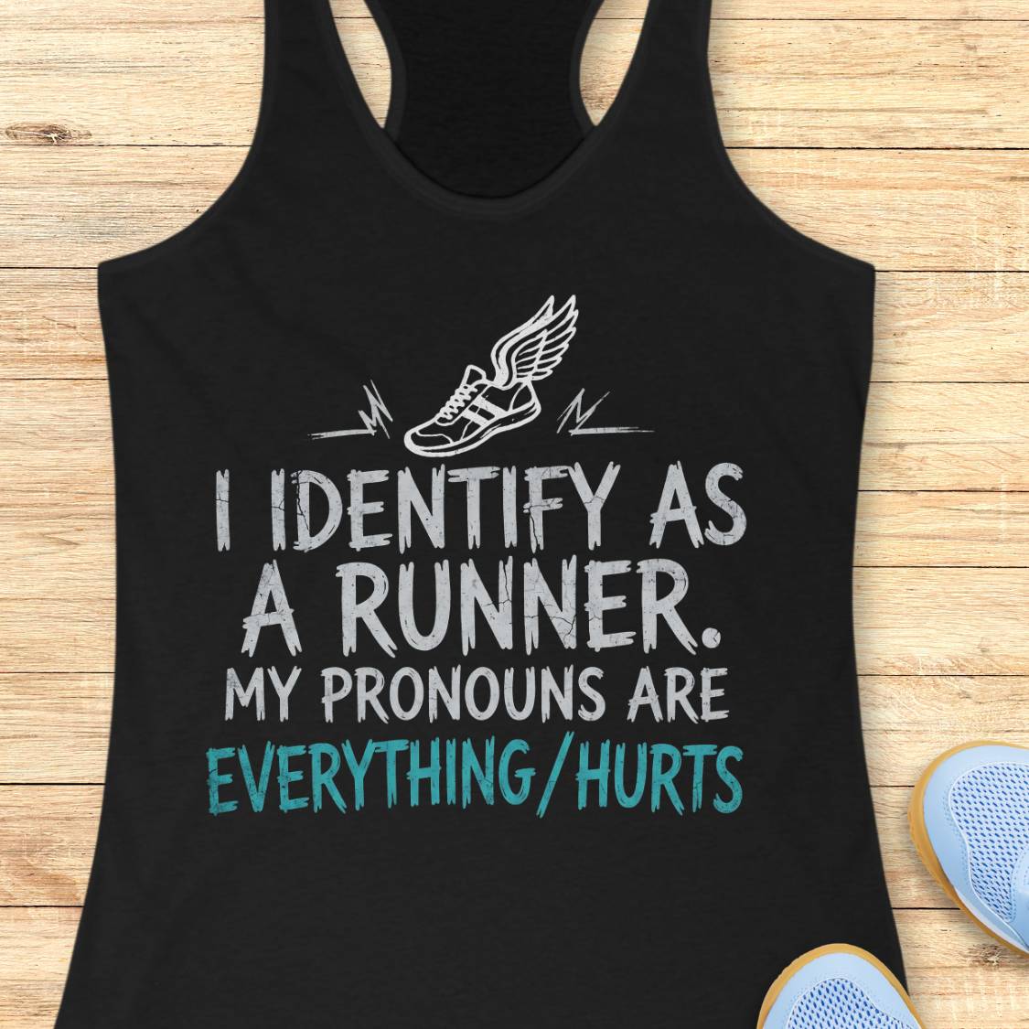 A black racerback tank top laid flat on a light wood background, featuring a white winged running shoe graphic and the text 'I IDENTIFY AS A RUNNER. MY PRONOUNS ARE EVERYTHING/HURTS' in white and teal.