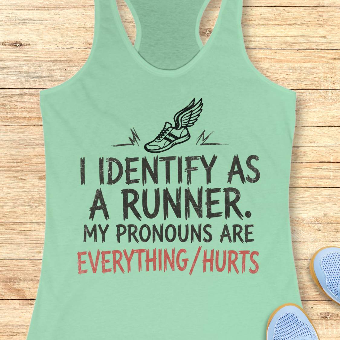 A mint green racerback tank top laid flat on a light wood background, featuring a black winged shoe graphic and the text 'I IDENTIFY AS A RUNNER. MY PRONOUNS ARE EVERYTHING/HURTS' in black and coral red lettering.