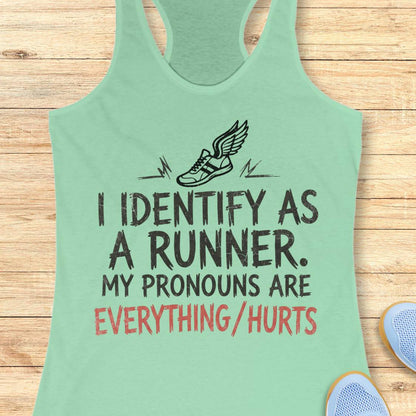 A mint green racerback tank top laid flat on a light wood background, featuring a black winged shoe graphic and the text 'I IDENTIFY AS A RUNNER. MY PRONOUNS ARE EVERYTHING/HURTS' in black and coral red lettering.