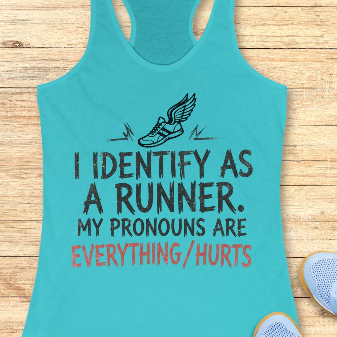 A blue racerback tank top laid flat on a light wood background, featuring a black winged shoe graphic and the text 'I IDENTIFY AS A RUNNER. MY PRONOUNS ARE EVERYTHING/HURTS' in black and coral red lettering.