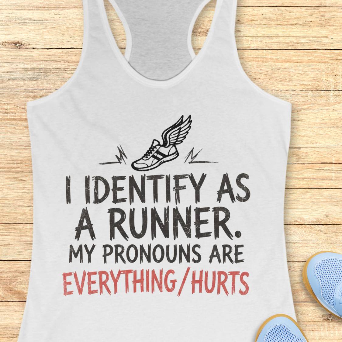 A white racerback tank top laid flat on a light wood background, featuring a black winged shoe graphic and the text 'I IDENTIFY AS A RUNNER. MY PRONOUNS ARE EVERYTHING/HURTS' in black and red lettering.