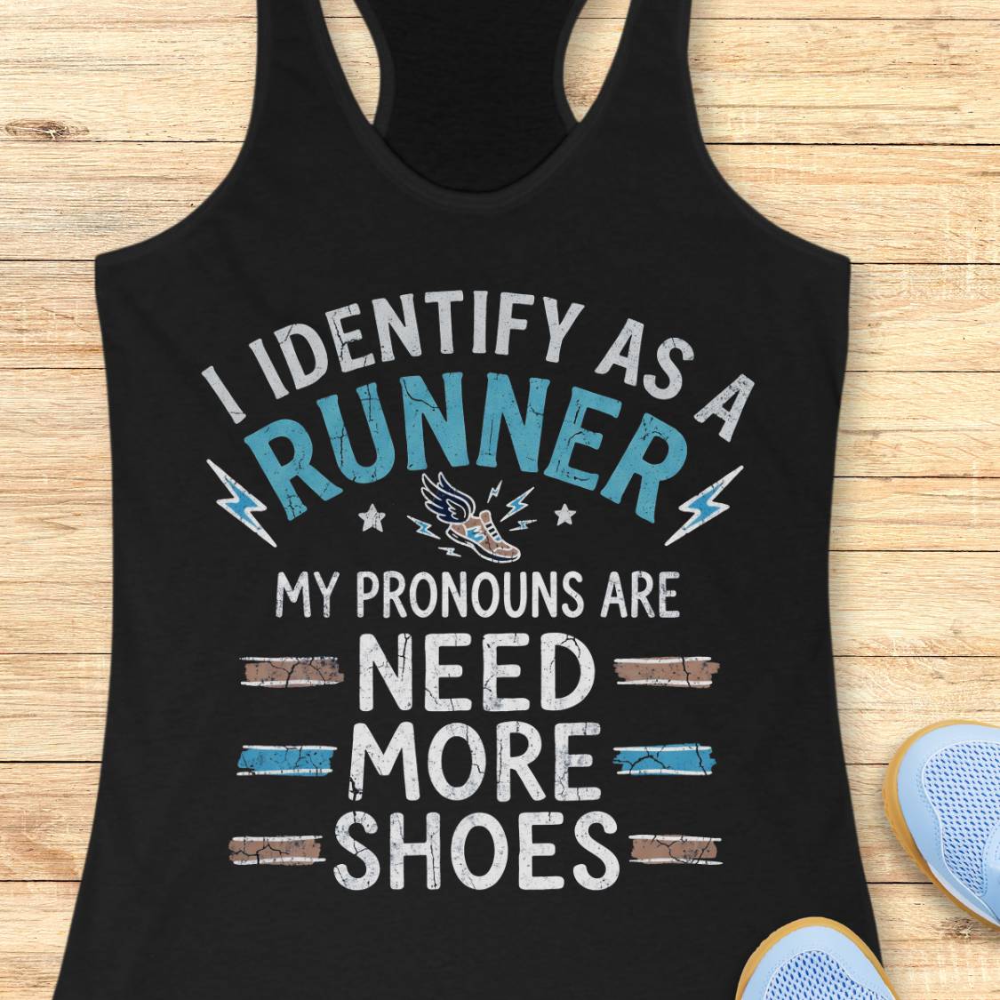A black racerback tank top laid flat on a light wood background, featuring a humorous graphic that reads 'I IDENTIFY AS A RUNNER MY PRONOUNS ARE NEED MORE SHOES' in distressed white and light blue lettering, with a small running shoe illustration. Partial blue sneakers appear at the bottom right as styling props.