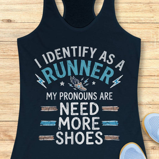 A navy blue racerback tank top laid flat on a light wood background, featuring a humorous graphic that reads 'I IDENTIFY AS A RUNNER MY PRONOUNS ARE NEED MORE SHOES' in white and light blue text with a small running shoe illustration.