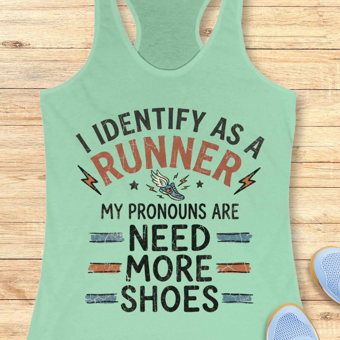 A mint green racerback tank top laid flat on a light wood background, featuring a humorous graphic print that reads 'I IDENTIFY AS A RUNNER MY PRONOUNS ARE NEED MORE SHOES' with small lightning bolts, stars, and a sneaker illustration.
