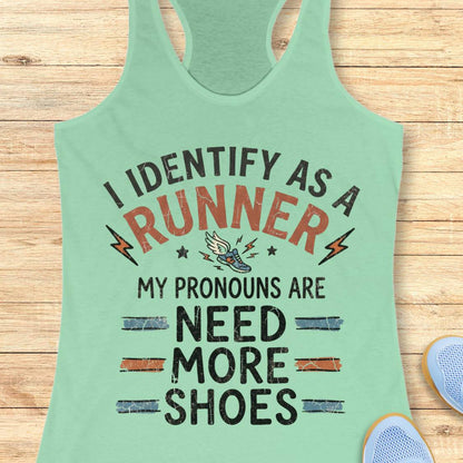 A mint green racerback tank top laid flat on a light wood background, featuring a humorous graphic print that reads 'I IDENTIFY AS A RUNNER MY PRONOUNS ARE NEED MORE SHOES' with small lightning bolts, stars, and a sneaker illustration.
