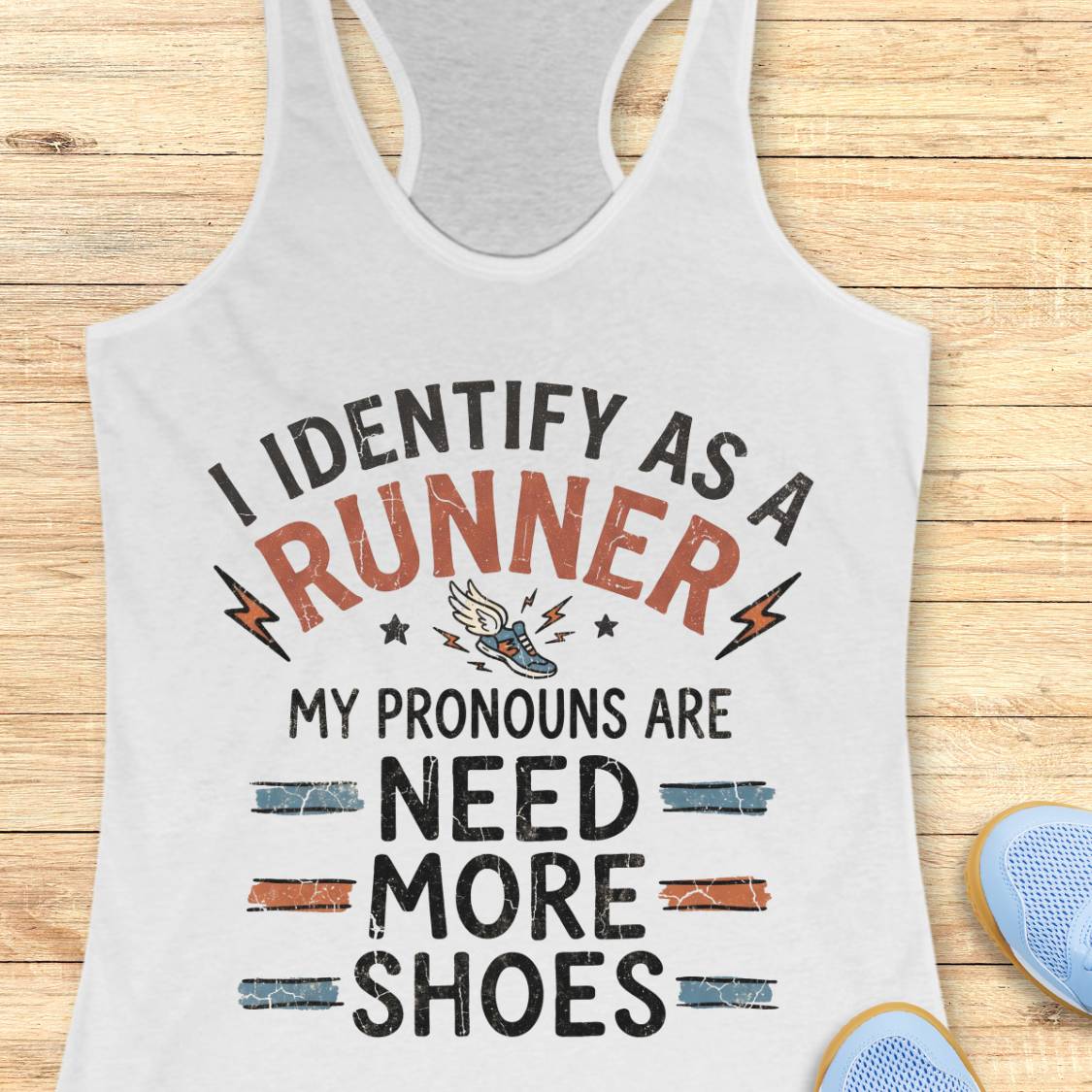 A white racerback tank top with a humorous graphic print reading 'I IDENTIFY AS A RUNNER MY PRONOUNS ARE NEED MORE SHOES' in black and orange lettering, displayed on a light wood background with blue sneakers partially visible in the corner.