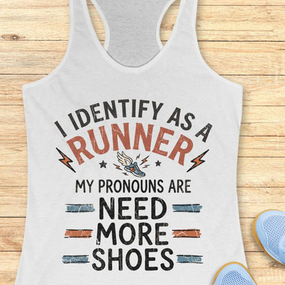 A white racerback tank top with a humorous graphic print reading 'I IDENTIFY AS A RUNNER MY PRONOUNS ARE NEED MORE SHOES' in black and orange lettering, displayed on a light wood background with blue sneakers partially visible in the corner.