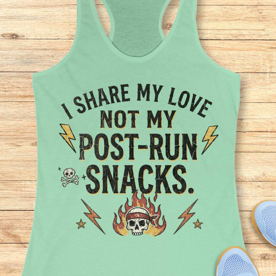 A mint green racerback tank top laid flat on a light wood background, featuring a distressed black graphic with the text 'I SHARE MY LOVE NOT MY POST-RUN SNACKS.' and illustrations of lightning bolts, a skull and crossbones, and a flaming skull wearing a headband.