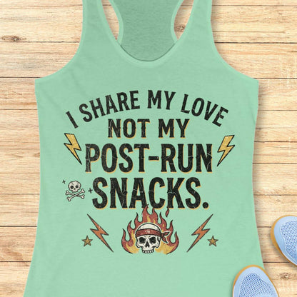 A mint green racerback tank top laid flat on a light wood background, featuring a distressed black graphic with the text 'I SHARE MY LOVE NOT MY POST-RUN SNACKS.' and illustrations of lightning bolts, a skull and crossbones, and a flaming skull wearing a headband.
