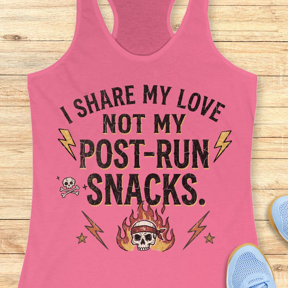 A pink racerback tank top laid flat on a light wood surface, featuring the printed phrase 'I SHARE MY LOVE NOT MY POST-RUN SNACKS.' in black with yellow lightning bolts, a skull with flames, and small decorative icons.