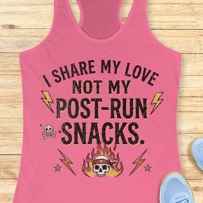 A pink racerback tank top laid flat on a light wood surface, featuring the printed phrase 'I SHARE MY LOVE NOT MY POST-RUN SNACKS.' in black with yellow lightning bolts, a skull with flames, and small decorative icons.