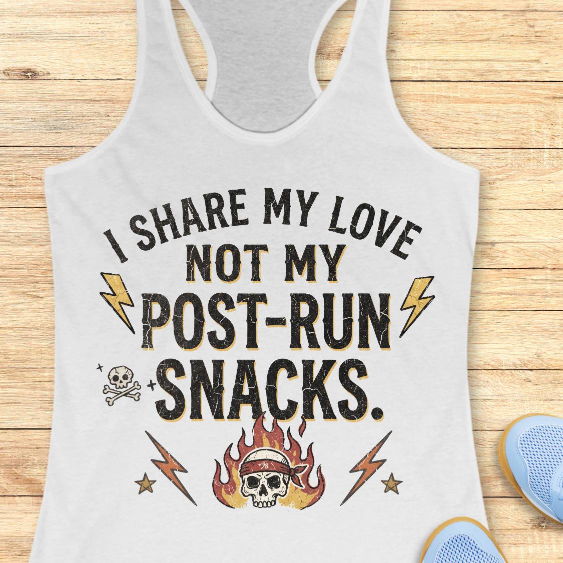 A white racerback tank top laid flat on a light wood plank background, featuring a distressed graphic that reads 'I SHARE MY LOVE NOT MY POST-RUN SNACKS.' with lightning bolts, stars, a skull, and a flaming skull illustration.