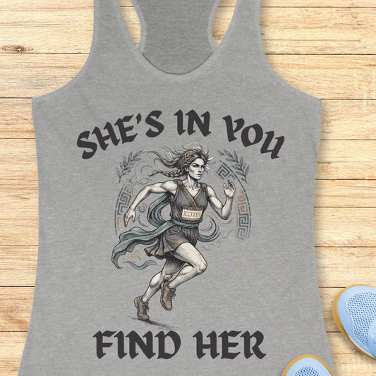 A heather gray racerback tank top featuring a graphic of a running woman with the text 'SHE'S IN YOU FIND HER' printed on the front, displayed flat on a light wood background.