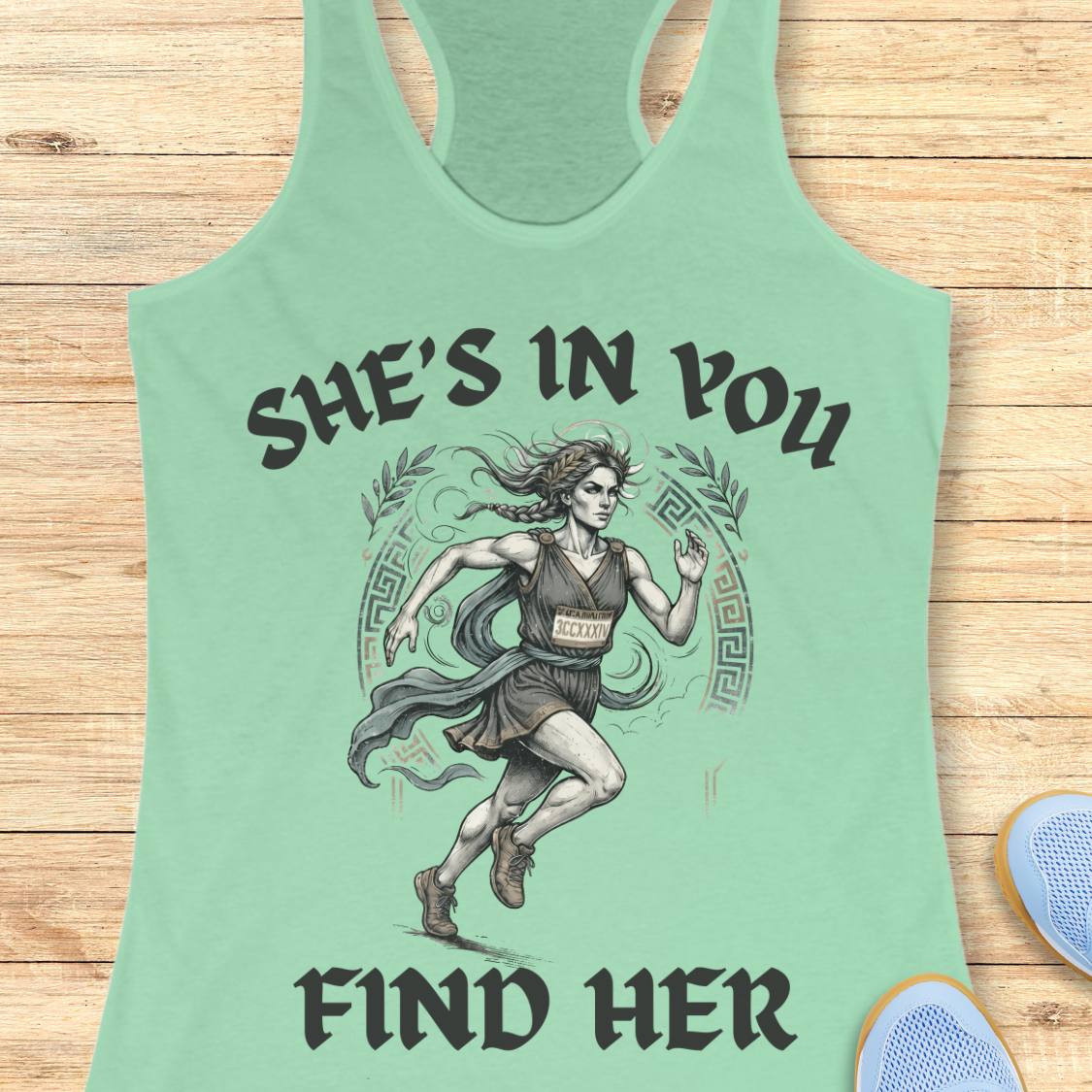 A mint racerback tank top featuring a graphic of a running woman with the text 'SHE'S IN YOU FIND HER' printed on the front, displayed flat on a light wood background.