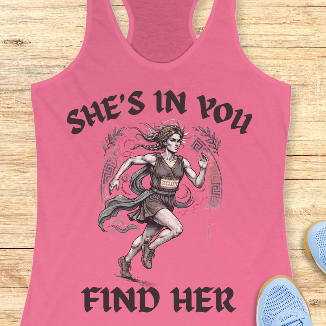 A pink racerback tank top laid flat on a light wood surface, featuring a graphic of a running woman with the text 'SHE'S IN YOU FIND HER' in black. Partial blue sneakers are visible at the bottom right as styling props.