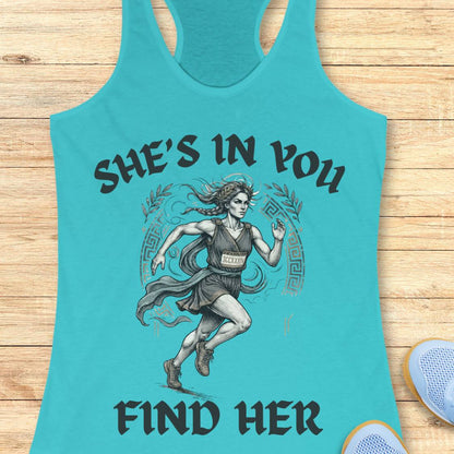 A blue racerback tank top featuring a graphic of a running woman with the text 'SHE'S IN YOU FIND HER' printed on the front, displayed flat on a light wood background.