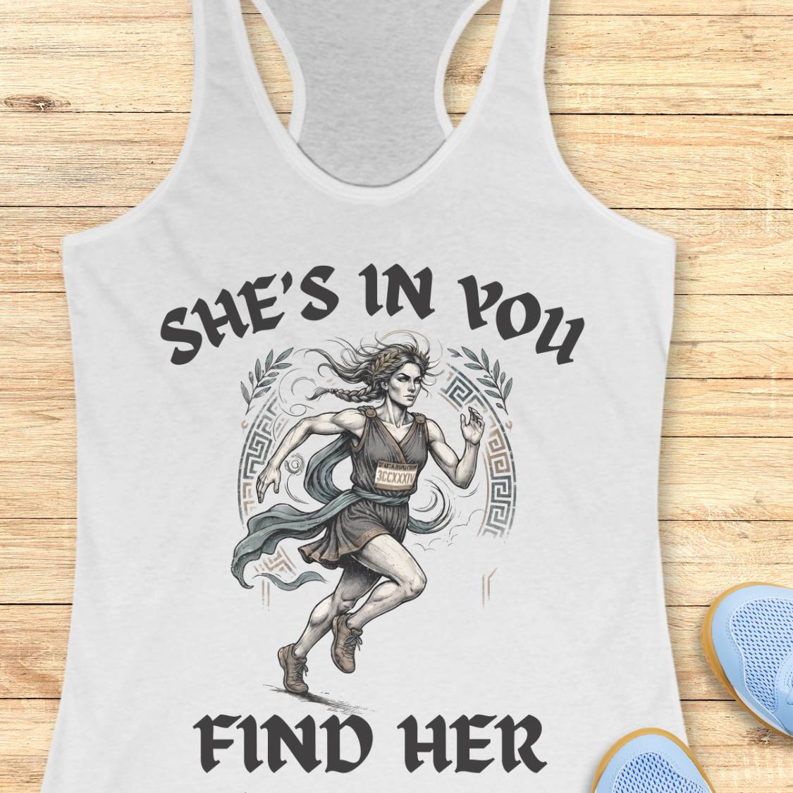 A white racerback tank top featuring a graphic of a running woman with the text 'SHE'S IN YOU FIND HER' printed on the front, displayed flat on a light wood background.