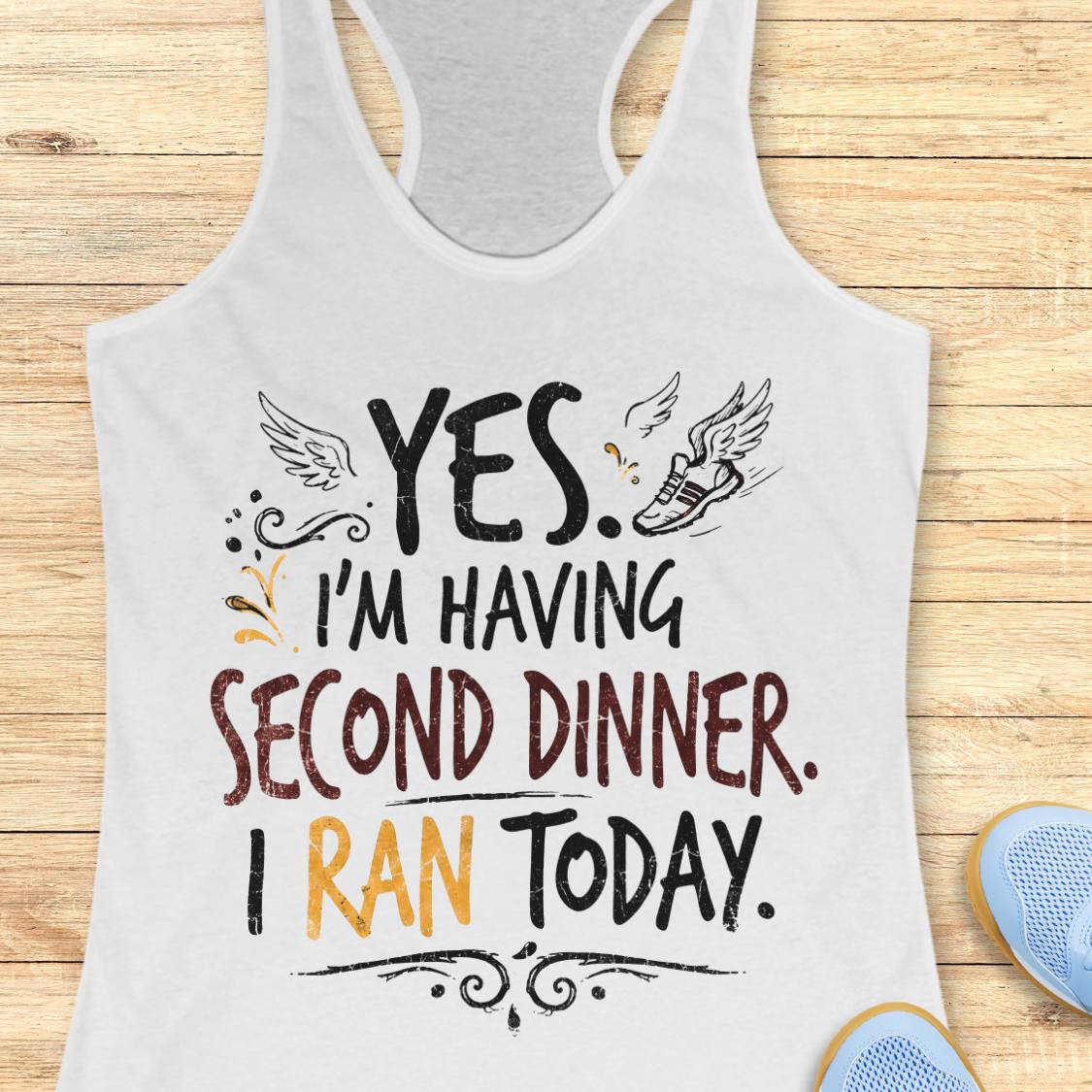 A white racerback tank top featuring  the text 'yes. I'm having second dinner. I ran today.' printed on the front, displayed flat on a light wood background.