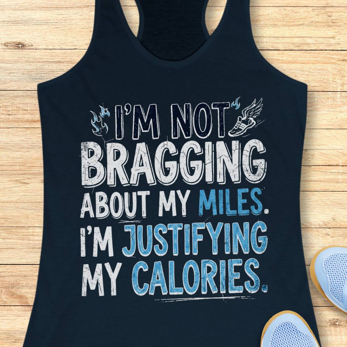 A  navy racerback tank top featuring  the text 'I'm not bragging about my miles. I'm justifying my calories' printed on the front, displayed flat on a light wood background.