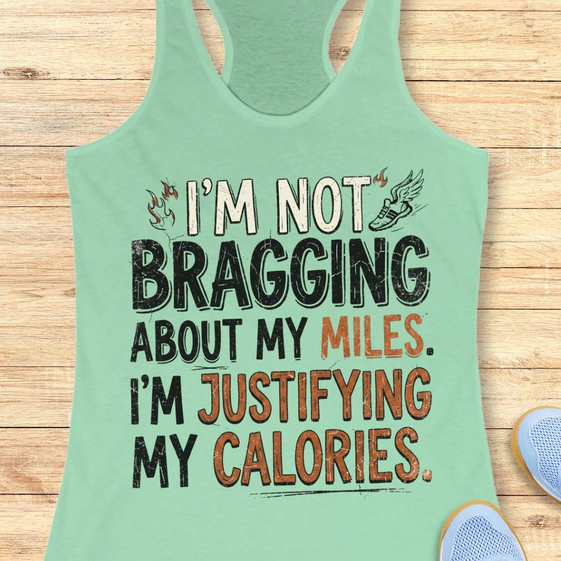 A  mint racerback tank top featuring  the text 'I'm not bragging about my miles. I'm justifying my calories' printed on the front, displayed flat on a light wood background.