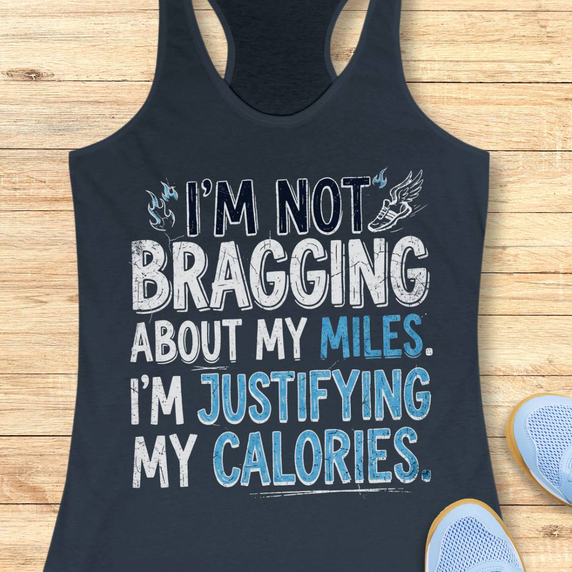 A  indigo racerback tank top featuring  the text 'I'm not bragging about my miles. I'm justifying my calories' printed on the front, displayed flat on a light wood background.
