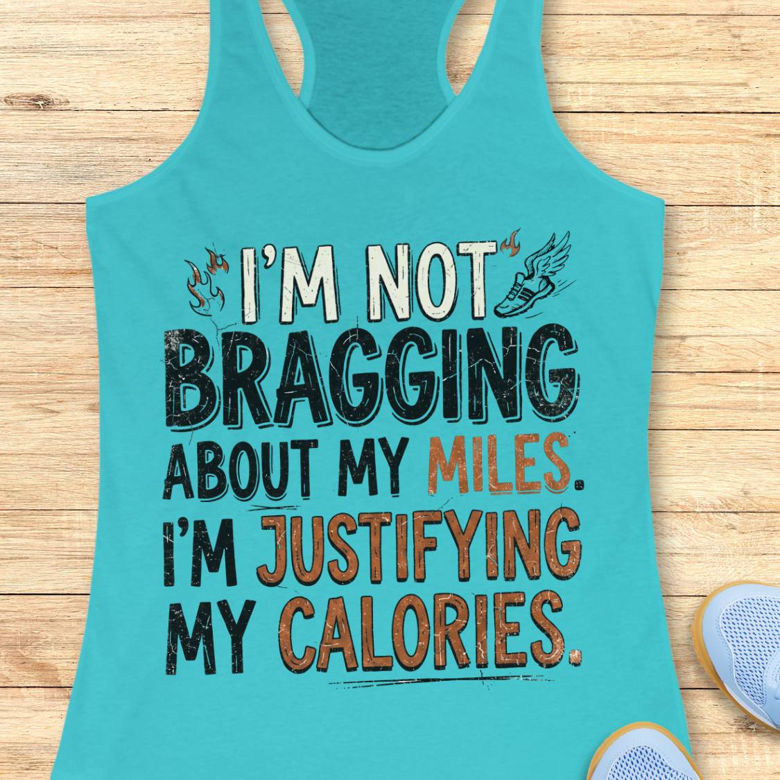 A  blue racerback tank top featuring  the text 'I'm not bragging about my miles. I'm justifying my calories' printed on the front, displayed flat on a light wood background.