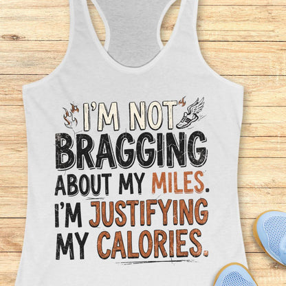 A  white racerback tank top featuring  the text 'I'm not bragging about my miles. I'm justifying my calories' printed on the front, displayed flat on a light wood background.