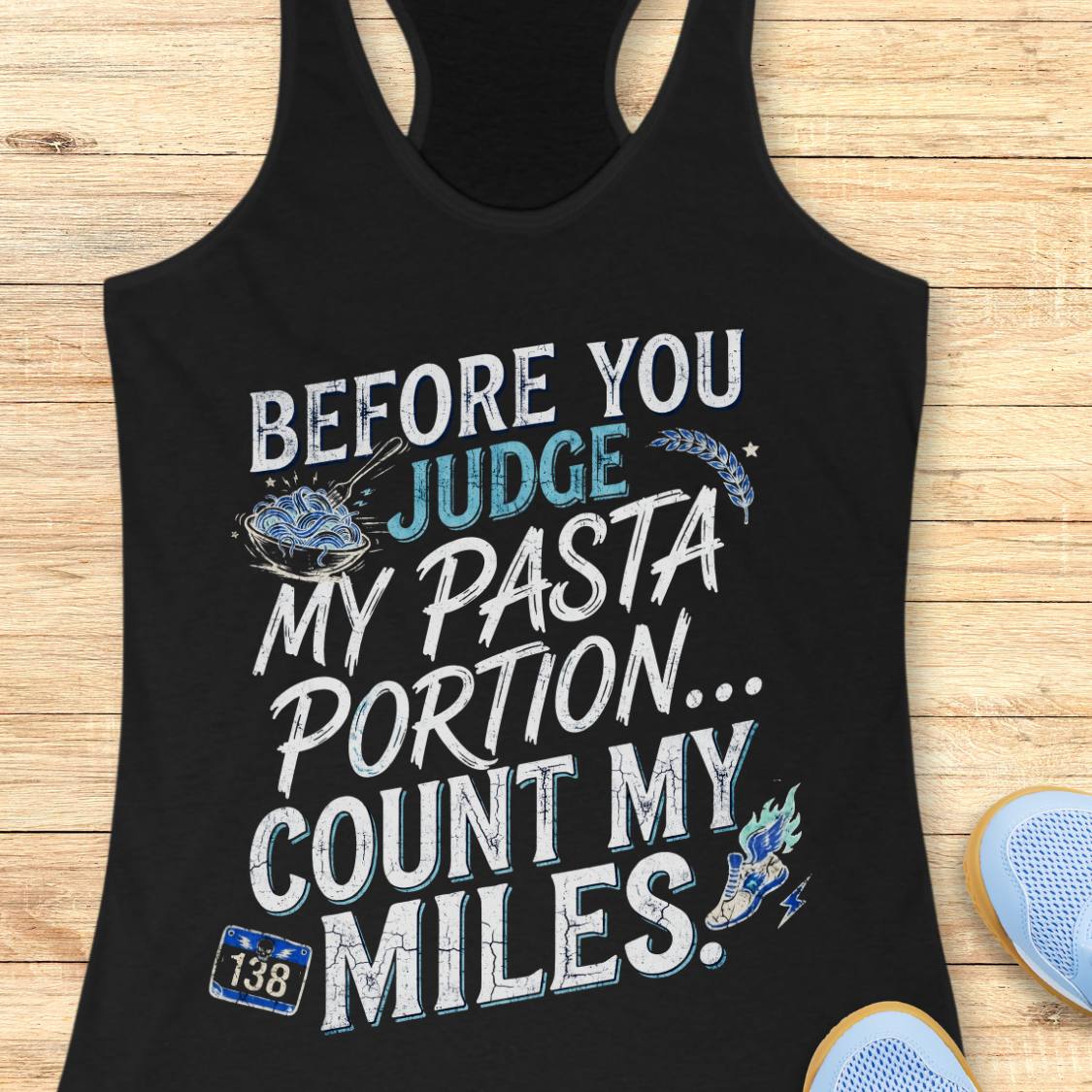 A black racerback tank top featuring  the text 'before you judge my pasta portion...count my miles ' printed on the front, displayed flat on a light wood background.