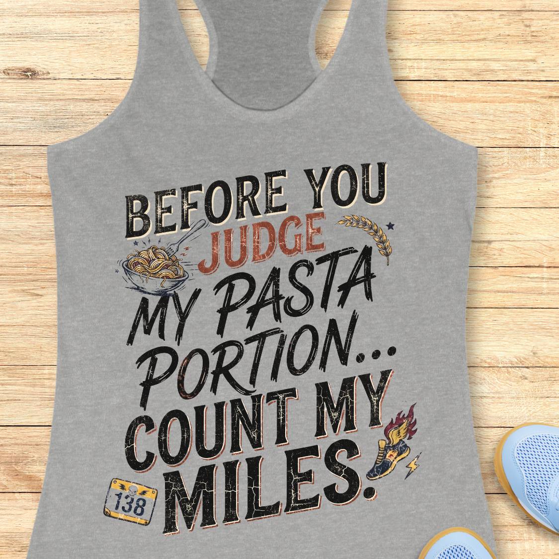 A grey racerback tank top featuring  the text 'before you judge my pasta portion...count my miles ' printed on the front, displayed flat on a light wood background.