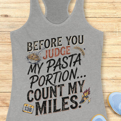 A grey racerback tank top featuring  the text 'before you judge my pasta portion...count my miles ' printed on the front, displayed flat on a light wood background.