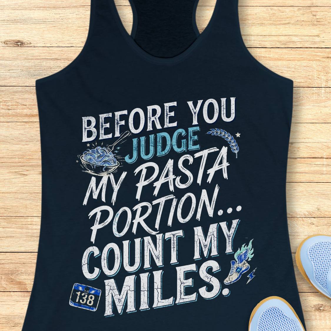 A midnight navy racerback tank top featuring  the text 'before you judge my pasta portion...count my miles ' printed on the front, displayed flat on a light wood background.