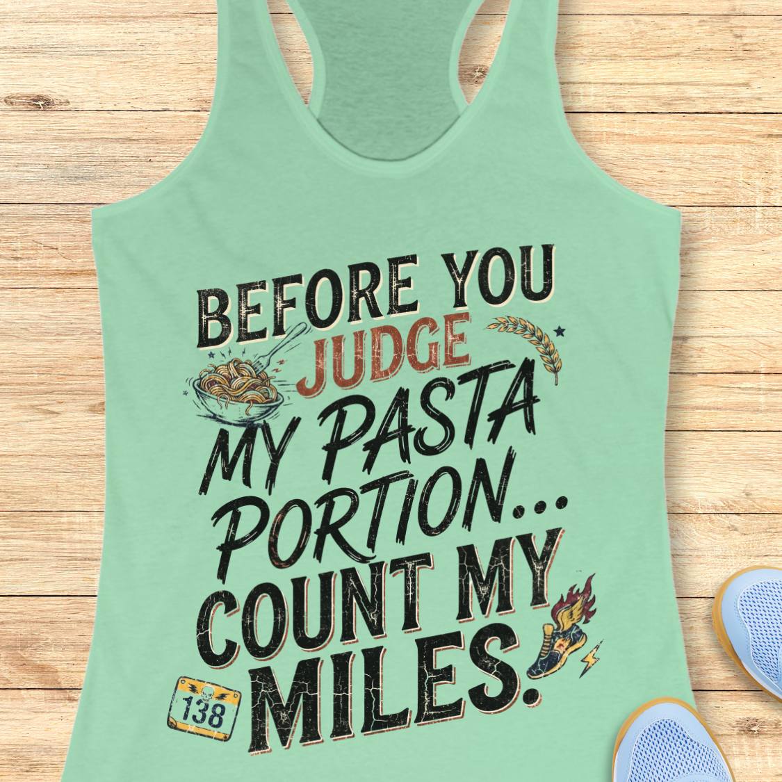 A mint racerback tank top featuring  the text 'before you judge my pasta portion...count my miles ' printed on the front, displayed flat on a light wood background.